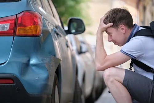 A man with liability only insurance looks worried as he crouches next to his vehicle after a fender bender.