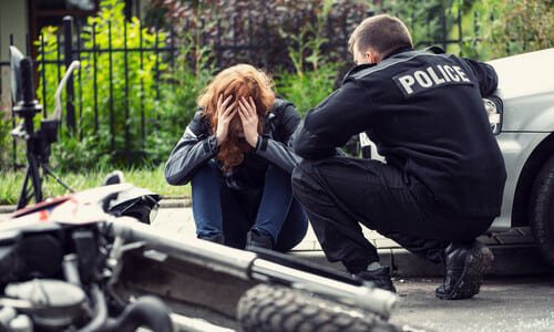 A woman sitting on a sidewalk crying, while a police officer tends to her, and with a motorcycle in the foreground.