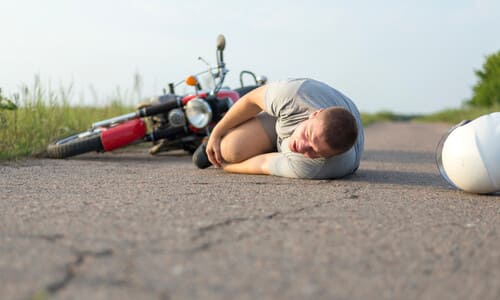 A man lying on the road while grasping his knee, and with his motorcycle on its side in the background.