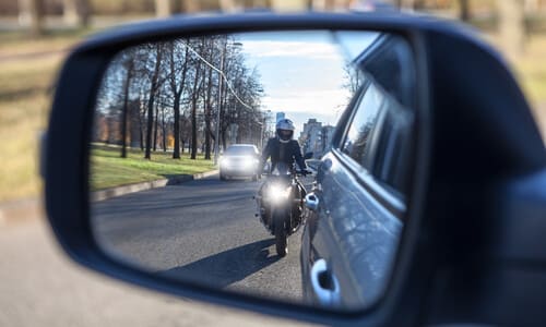 A car's left side view mirror reflecting a motorcycle attempting to overtake.