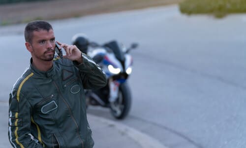 A male motorcyclist on the phone at the side of the road, with hsi bike in the background.