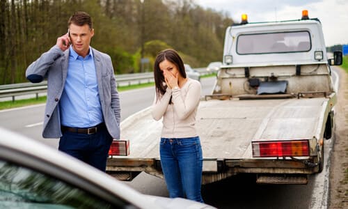 A man and woman on a phone call after their car was involved in an accident with a truck in the background.